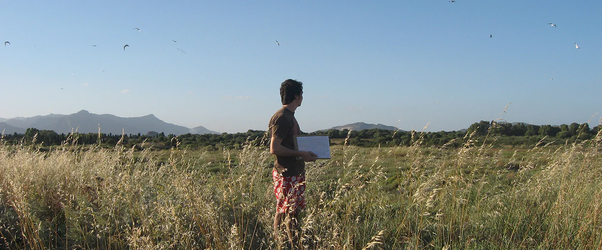 1_A Ricercatore osserva il volo di uccelli in una zona umida, tenendo una griglia per il monitoraggio dell’avifauna, circondato da vegetazione spontanea e montagne all’orizzonte.