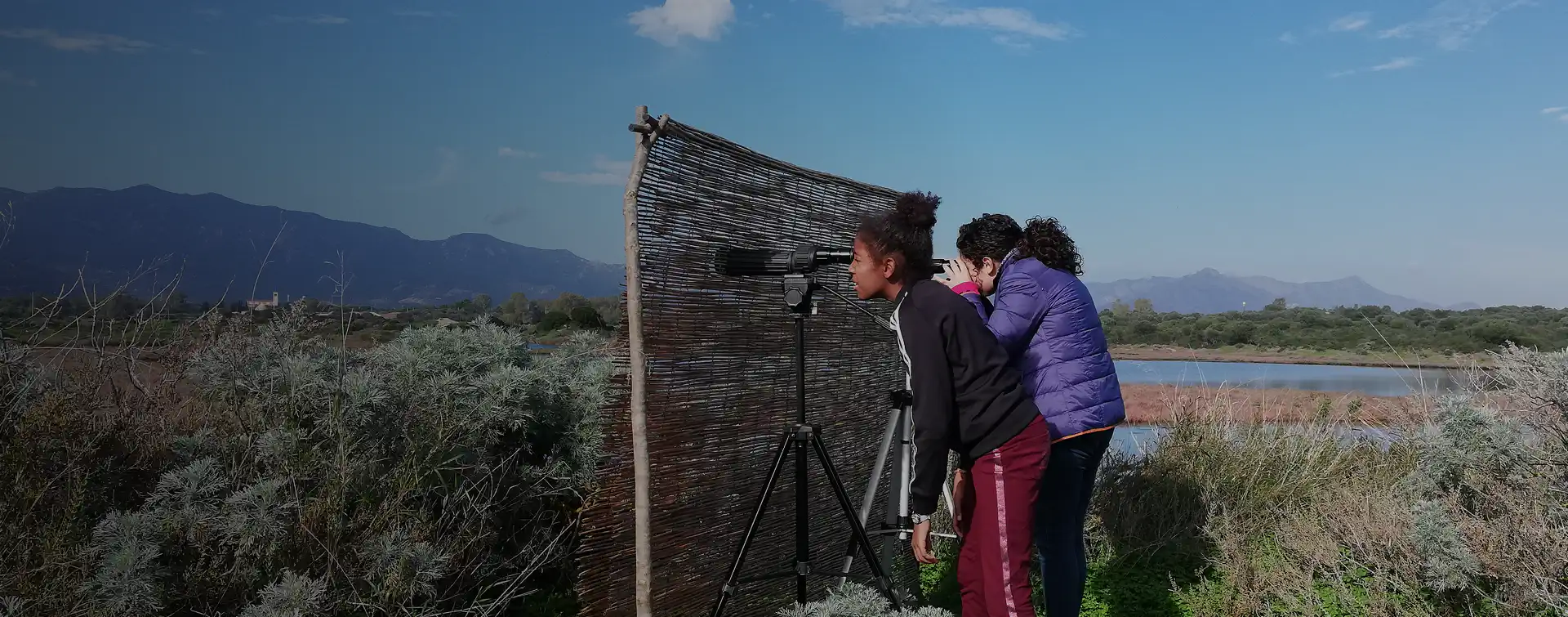 Due ragazze osservano gli uccelli attraverso un binocolo posizionato su un treppiede, nascosto dietro una barriera mimetica nella zona umida della Laguna di Nora, con lo sfondo delle montagne e della vegetazione mediterranea.