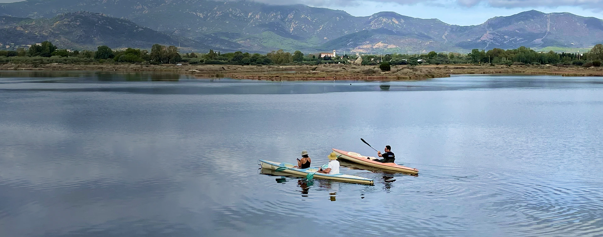 Tre persone in canoa esplorano le acque tranquille della Laguna di Nora, circondate da natura incontaminata e con le montagne sullo sfondo.
