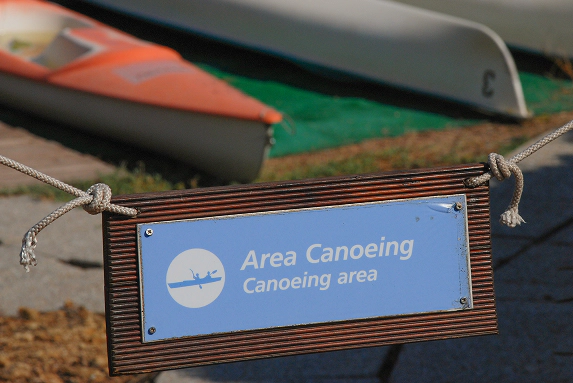1_C Cartello in legno con scritta “Area Canoeing” davanti a canoe posate a terra in un’area dedicata allo sport acquatico.