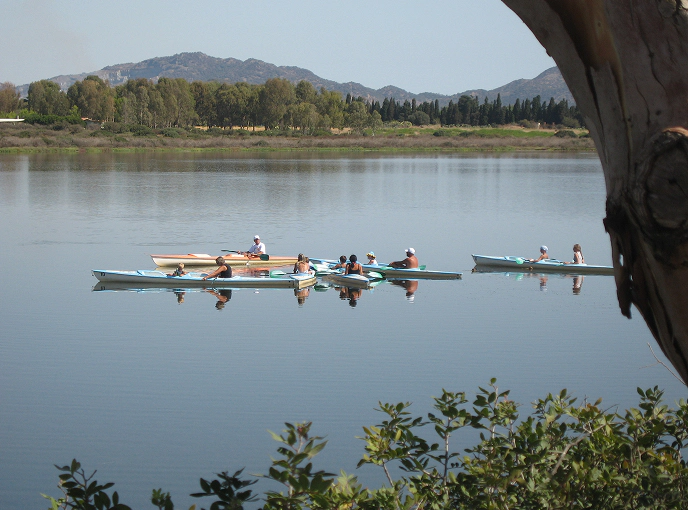 1_D Tre canoe con sportivi in allenamento solcano le acque tranquille di uno stagno circondato dalla vegetazione, con sfondo di montagne e pini tipici del paesaggio sardo.