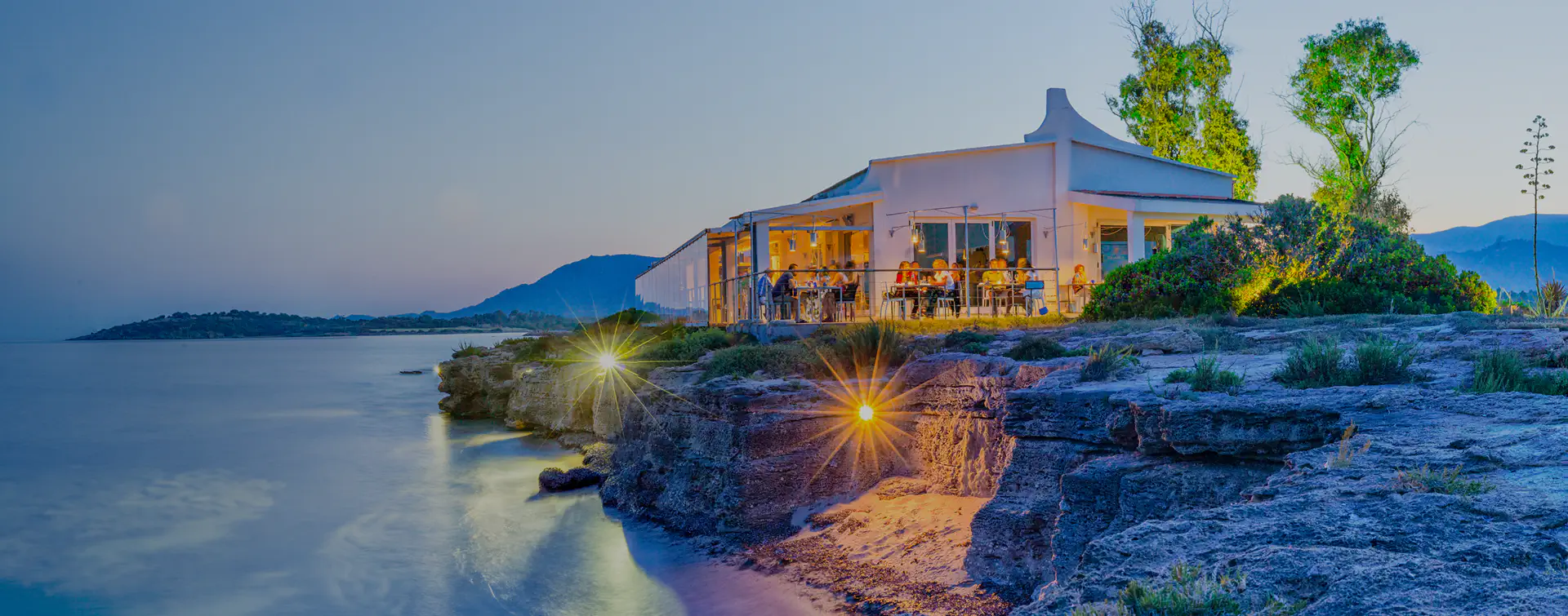 Vista panoramica al tramonto sul ristorante fronte mare della Laguna di Nora, con luci calde, scogliera rocciosa, vegetazione mediterranea e montagne sullo sfondo.
