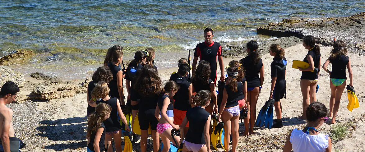 2_A Gruppo di bambini e bambine con pinne e mute corte ascolta un istruttore su una spiaggia rocciosa della Sardegna, preparandosi a un’attività di snorkeling educativo.