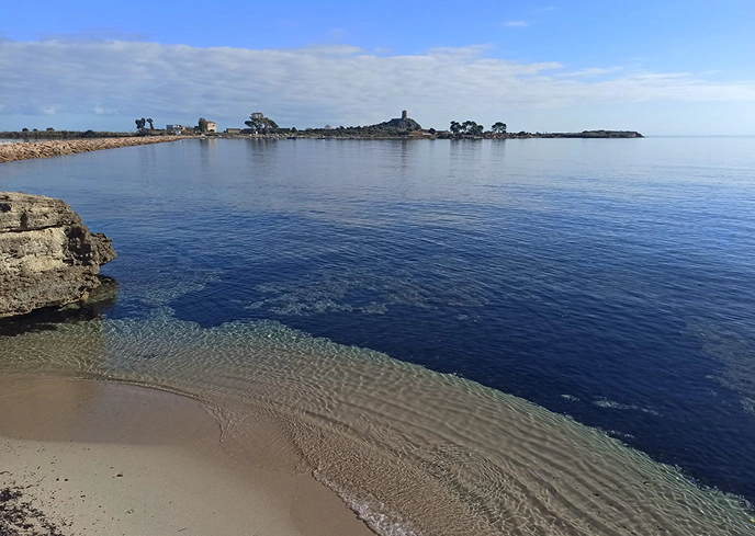 2_B Tratto costiero con spiaggia sabbiosa e mare cristallino, sullo sfondo l’area archeologica di Nora in Sardegna con la torre spagnola visibile all’orizzonte.