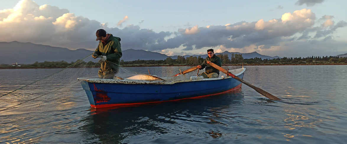 4_A Due pescatori in barca mentre recuperano una rete da pesca al tramonto, circondati da acque calme e montagne sullo sfondo.