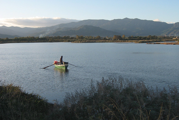 4_C Pescatore in barca a remi nella Laguna di Nora al tramonto, circondato da acqua calma e natura incontaminata con le montagne sullo sfondo.