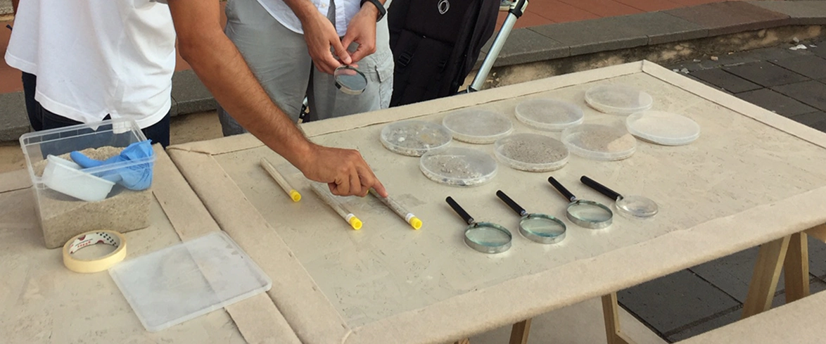 Tavolo didattico allestito con sabbia, lenti d’ingrandimento e piastre per analizzare la presenza di microplastiche nei campioni, durante un laboratorio educativo sull'inquinamento marino.