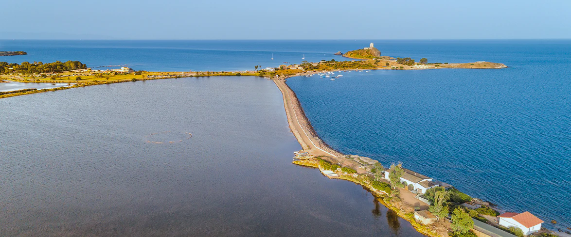 Veduta aerea della penisola di Nora in Sardegna, collegata da un sottile istmo tra mare aperto e laguna costiera, con torri storiche, acque cristalline e vegetazione mediterranea.