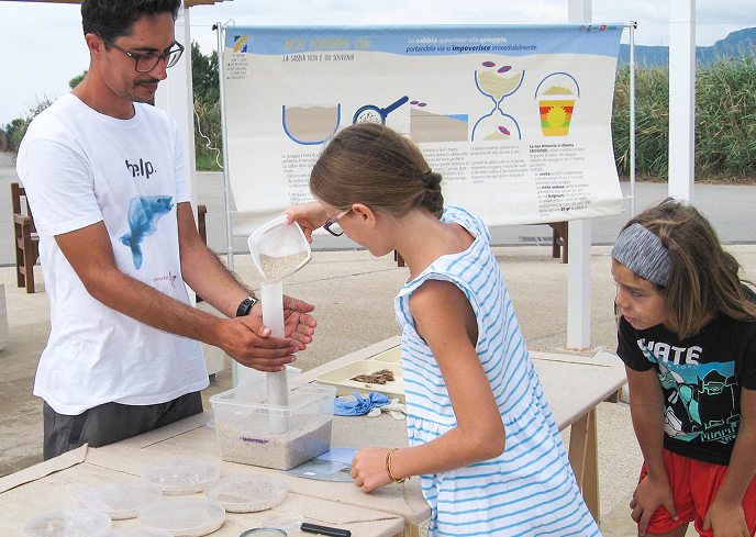 Attività educativa all'aperto con bambini che imparano a filtrare la sabbia in un laboratorio scientifico sul tema della microplastica e dell'inquinamento marino.