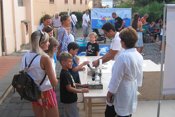 Bambini e famiglie partecipano a un laboratorio scientifico all'aperto con microscopi durante un evento educativo su ambiente e biodiversità.