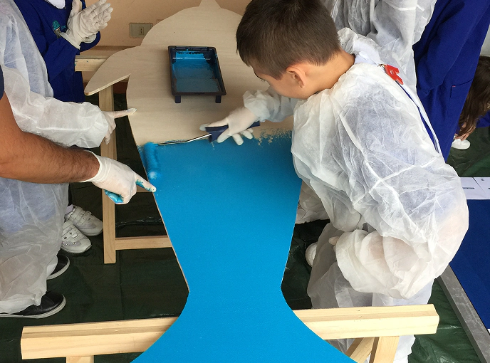 Bambino con tuta protettiva bianca dipinge di blu la sagoma di un pesce durante un laboratorio creativo ambientale per bambini.