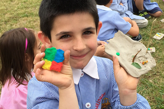 Bambino sorride mostrando due creazioni artistiche fatte a mano: una con pasta modellabile colorata e l’altra con elementi naturali su argilla, durante un'attività di educazione ambientale all'aperto.