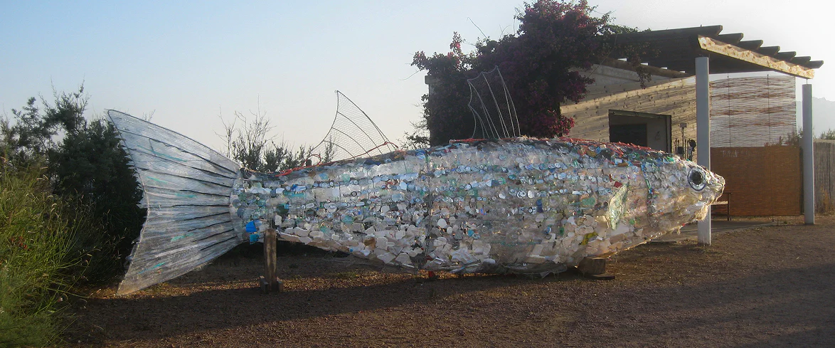 Grande scultura a forma di pesce realizzata con plastica riciclata e materiali di recupero, posizionata all’esterno di un edificio in legno come installazione di sensibilizzazione sull’inquinamento marino.