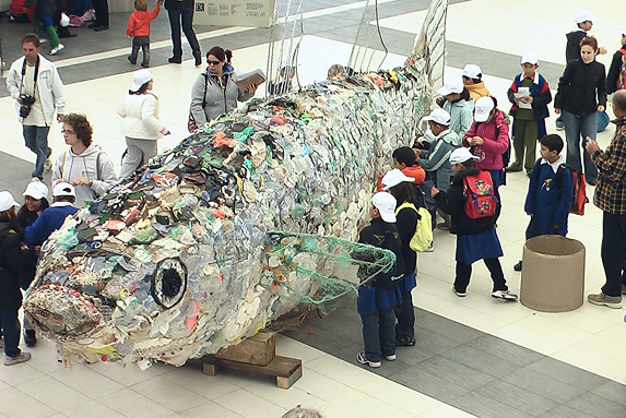 Scultura gigante a forma di pesce realizzata con rifiuti di plastica raccolti in mare, esposta in una piazza interna affollata da bambini con cappellini e zaini, coinvolti in un’attività educativa sul riciclo e l’inquinamento marino.