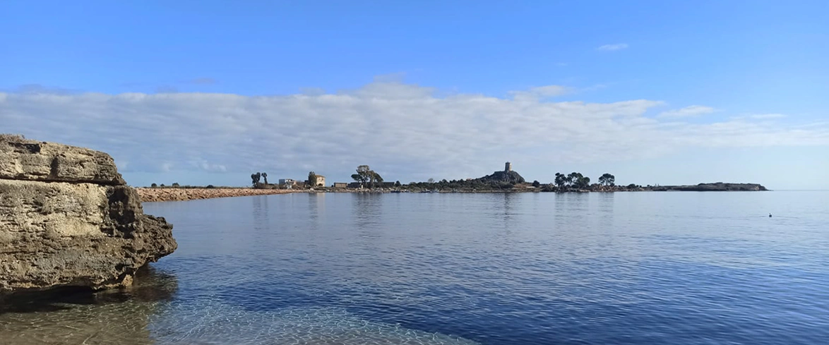 Panorama costiero con acque limpide e calme davanti alla penisola di Nora, in Sardegna, con scogliere rocciose in primo piano e la torre aragonese sullo sfondo sotto un cielo sereno.