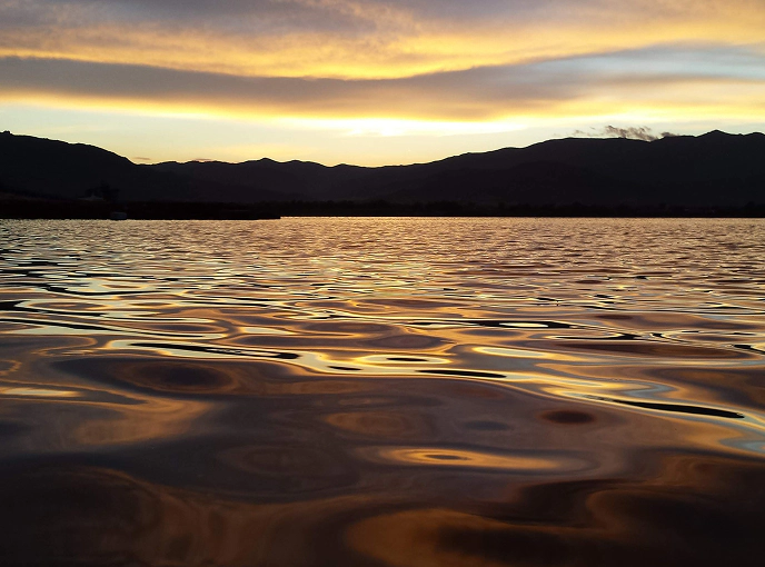 Tramonto su una laguna costiera con riflessi dorati e viola sull’acqua increspata, circondata da profili montuosi in controluce.